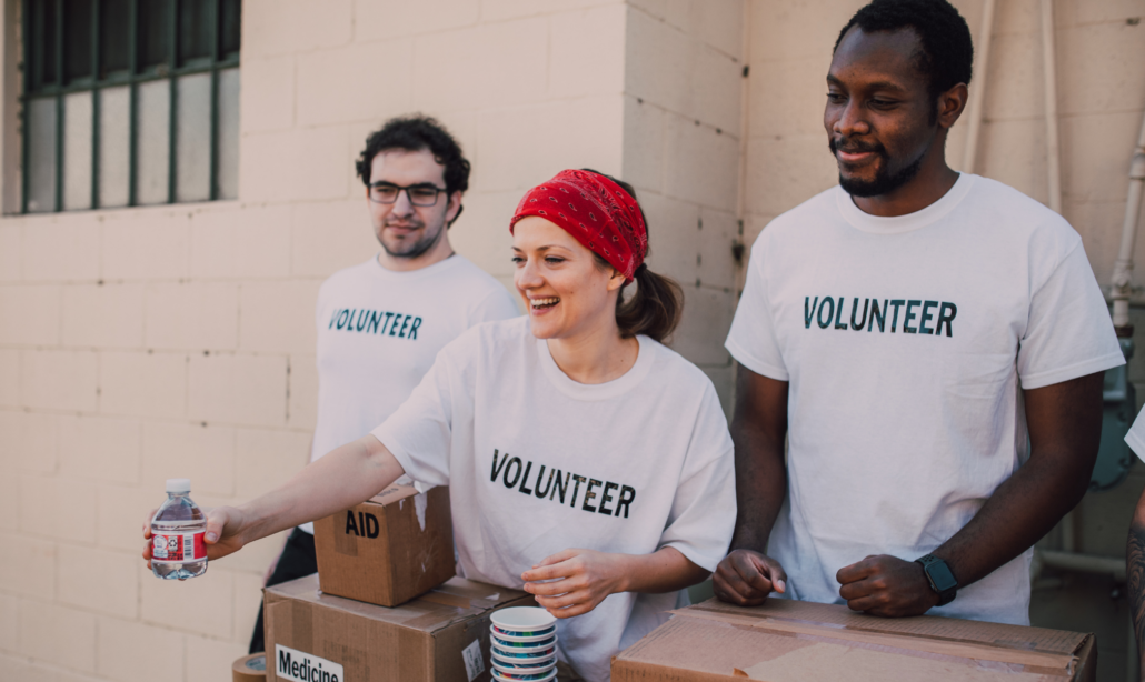 Drei Freiwillige in T-Shirts mit der Aufschrift "Volunteer" verteilen Hilfsgüter, darunter Wasserflaschen und Kartons mit medizinischer Versorgung.